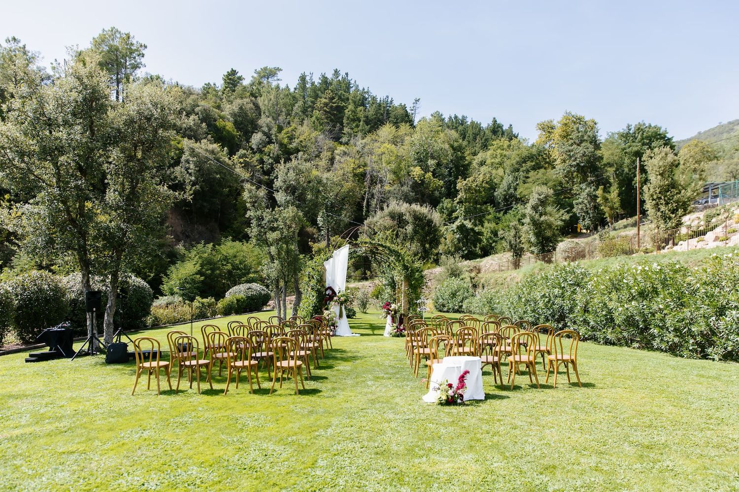 Outdoor wedding ceremony setup with wooden chairs on green lawn surrounded by lush trees and floral arch backdrop.