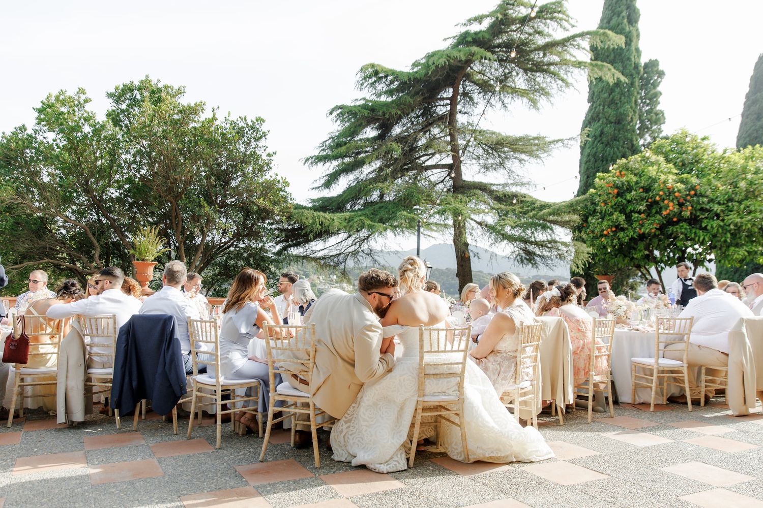 Outdoor wedding reception with guests seated at elegant tables surrounded by lush trees and a scenic backdrop.