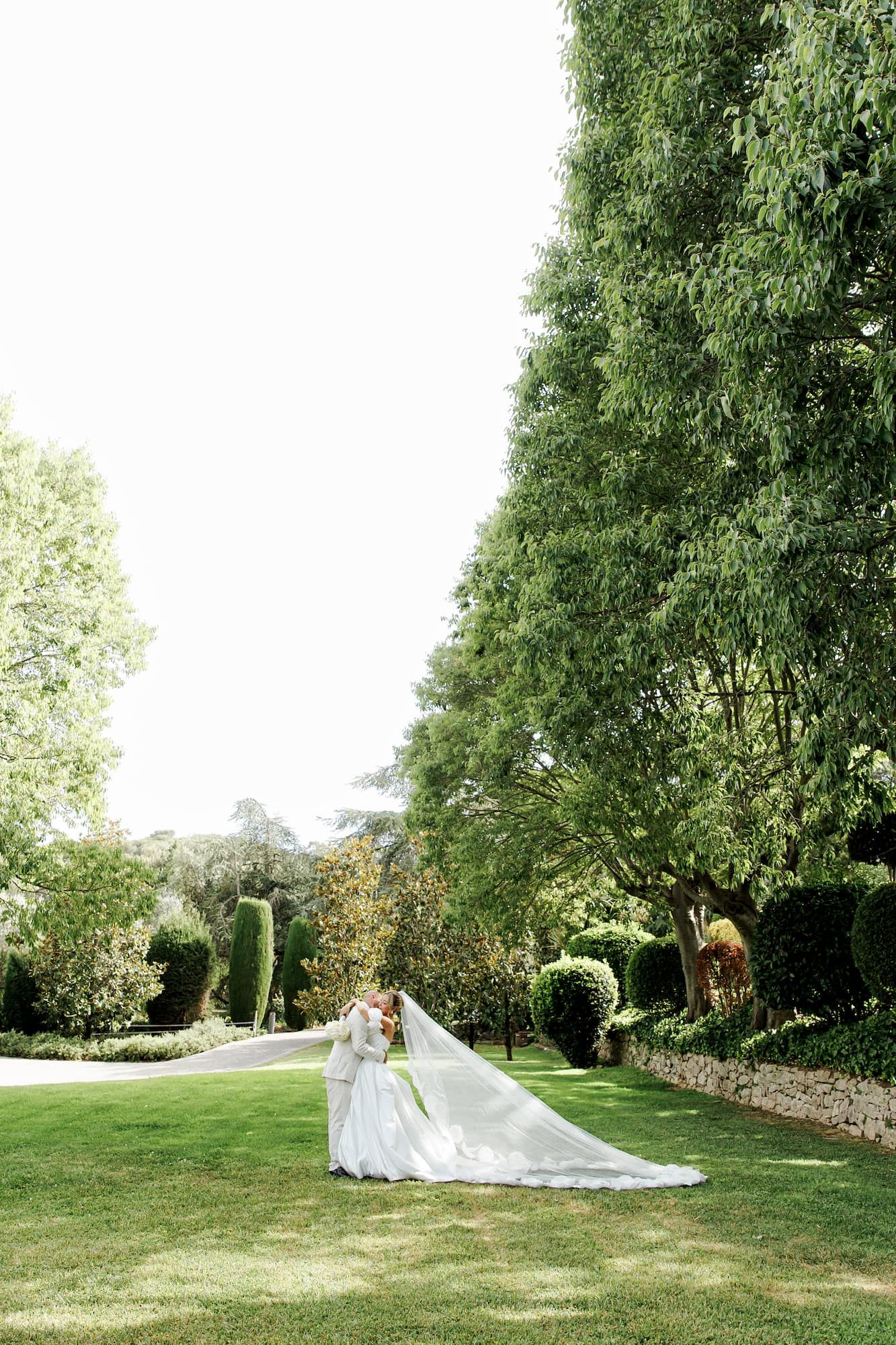 Bride in a flowing white gown with long veil billowing in a lush green garden surrounded by tall trees.