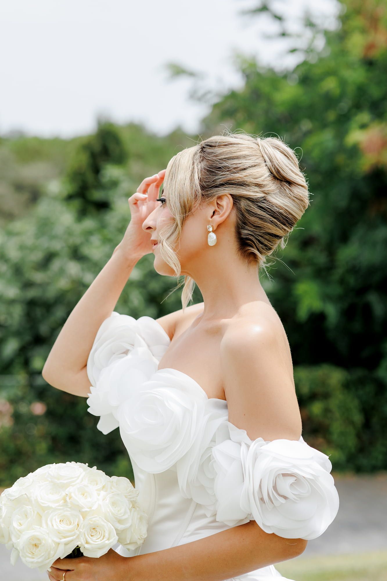 Elegant bride in off-shoulder white gown with rose details holds bouquet of white roses outdoors.