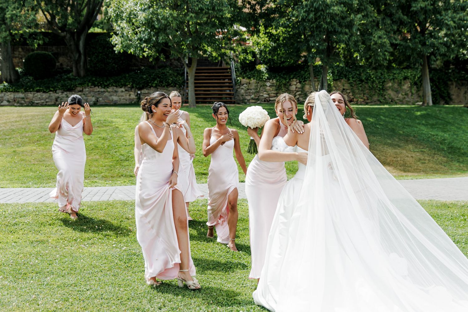 Bride in white gown and veil walks with bridesmaids in blush dresses on green lawn at outdoor wedding.