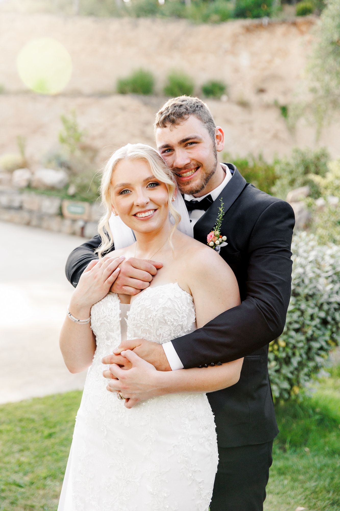 The groom is hugging the bride from the back and they look at the camera