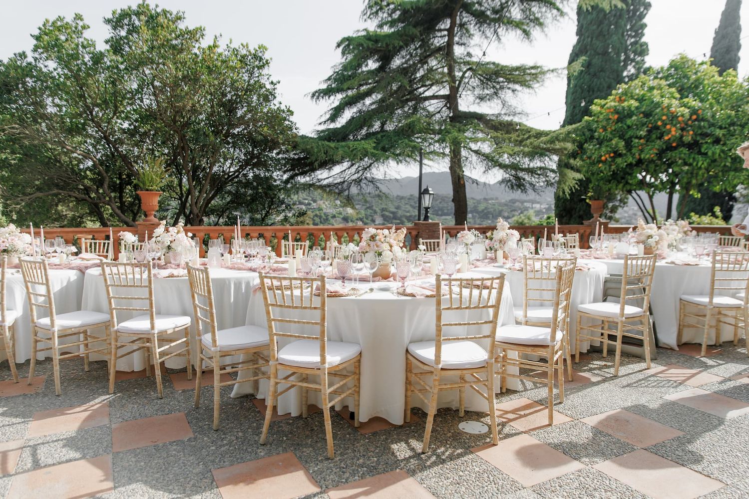 Elegant outdoor wedding reception with gold Chiavari chairs, white round tables, and lush greenery under a clear blue sky.