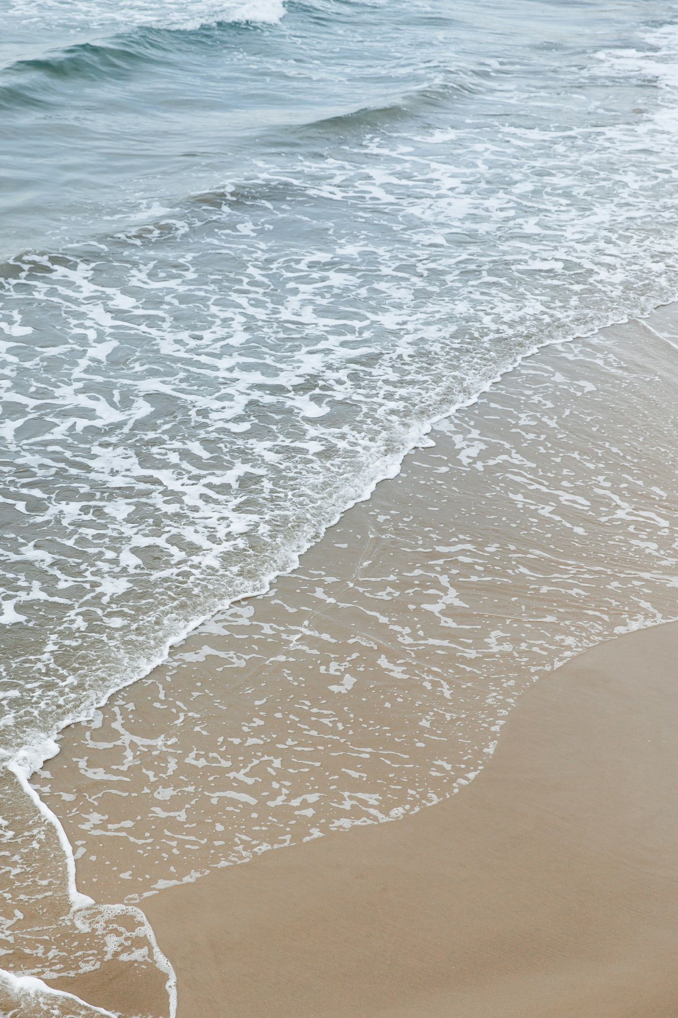 Ocean waves gently rolling onto a sandy beach with white foam creating intricate patterns along the shoreline.