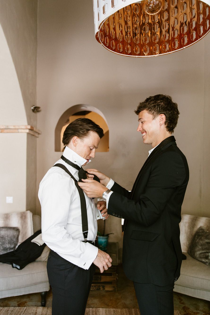 Groom getting his bow tie adjusted by smiling best man in black tuxedos before wedding ceremony.