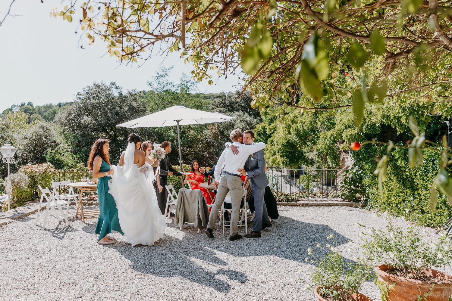 Wedding guests gather outdoors in a sunlit garden, celebrating with the bride and groom on a gravel terrace.
