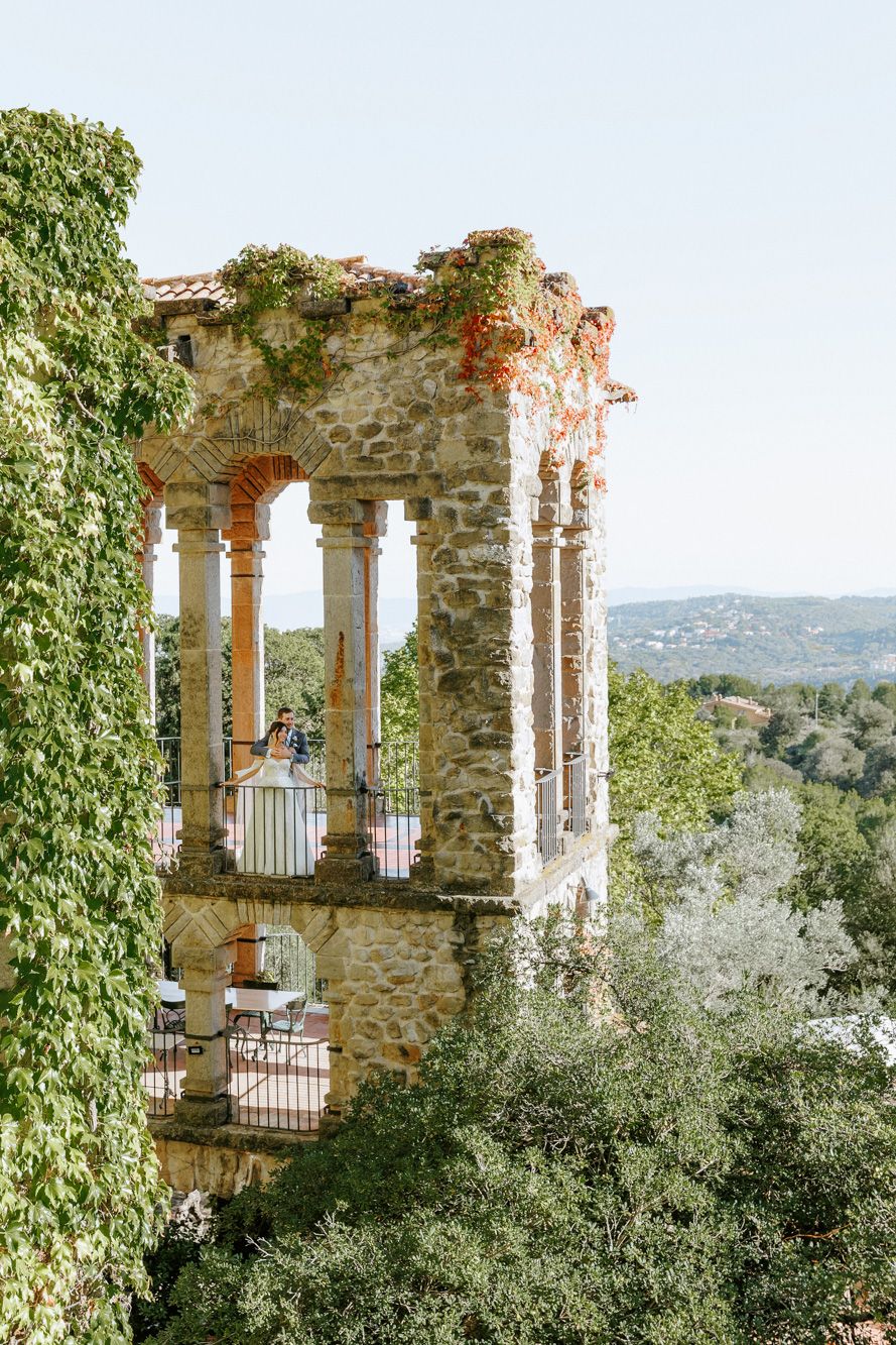 Ancient stone tower with ivy-covered walls, arched columns, and La Baronia balcony overlooking lush green hills.