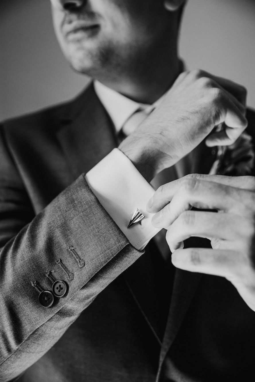 Black and white photo of a groom in a suit fastening his cufflinks while adjusting his tie on his wedding day.