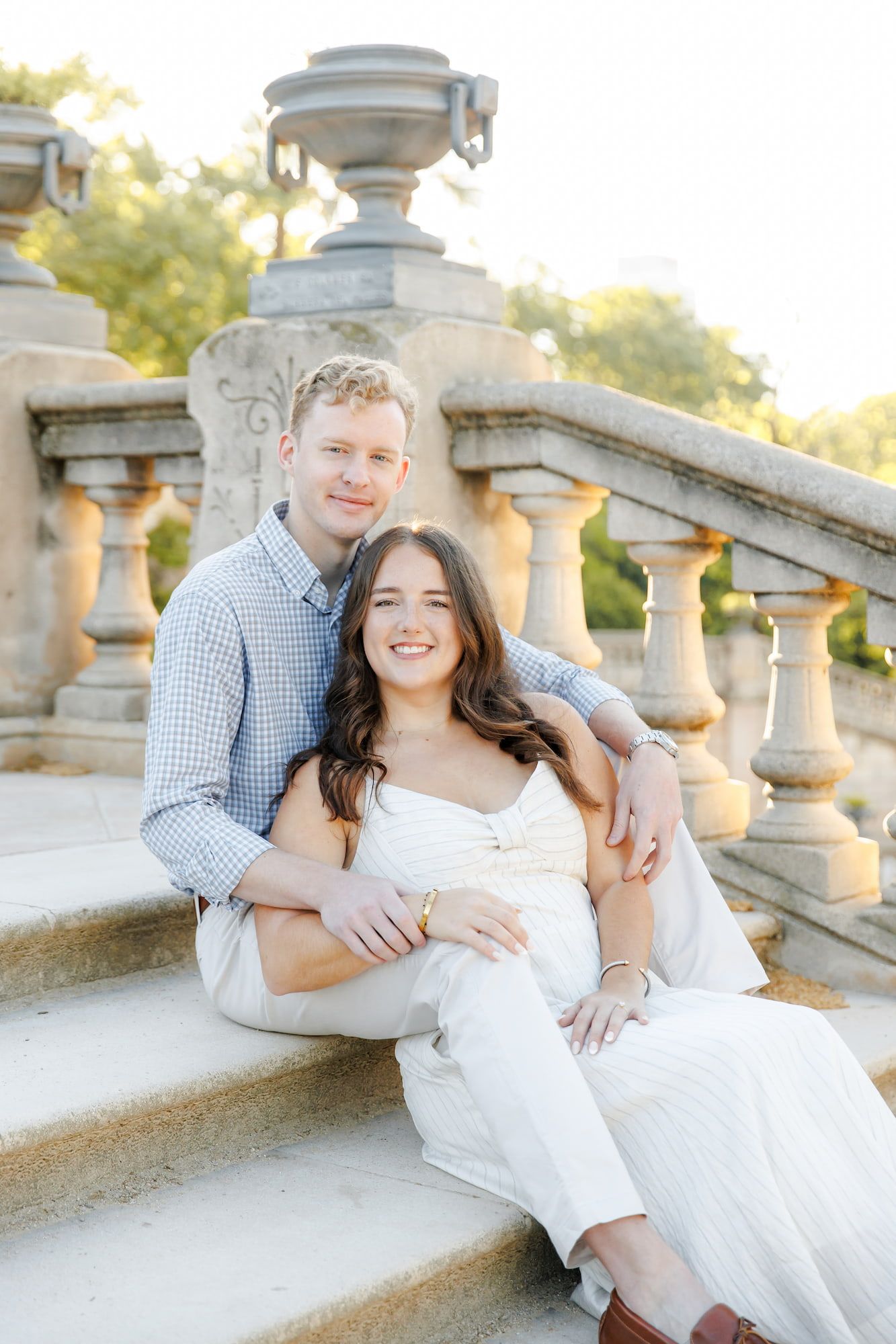 Couple sitting on ornate stone steps smiling, man in blue shirt embracing woman in white dress at golden hour.