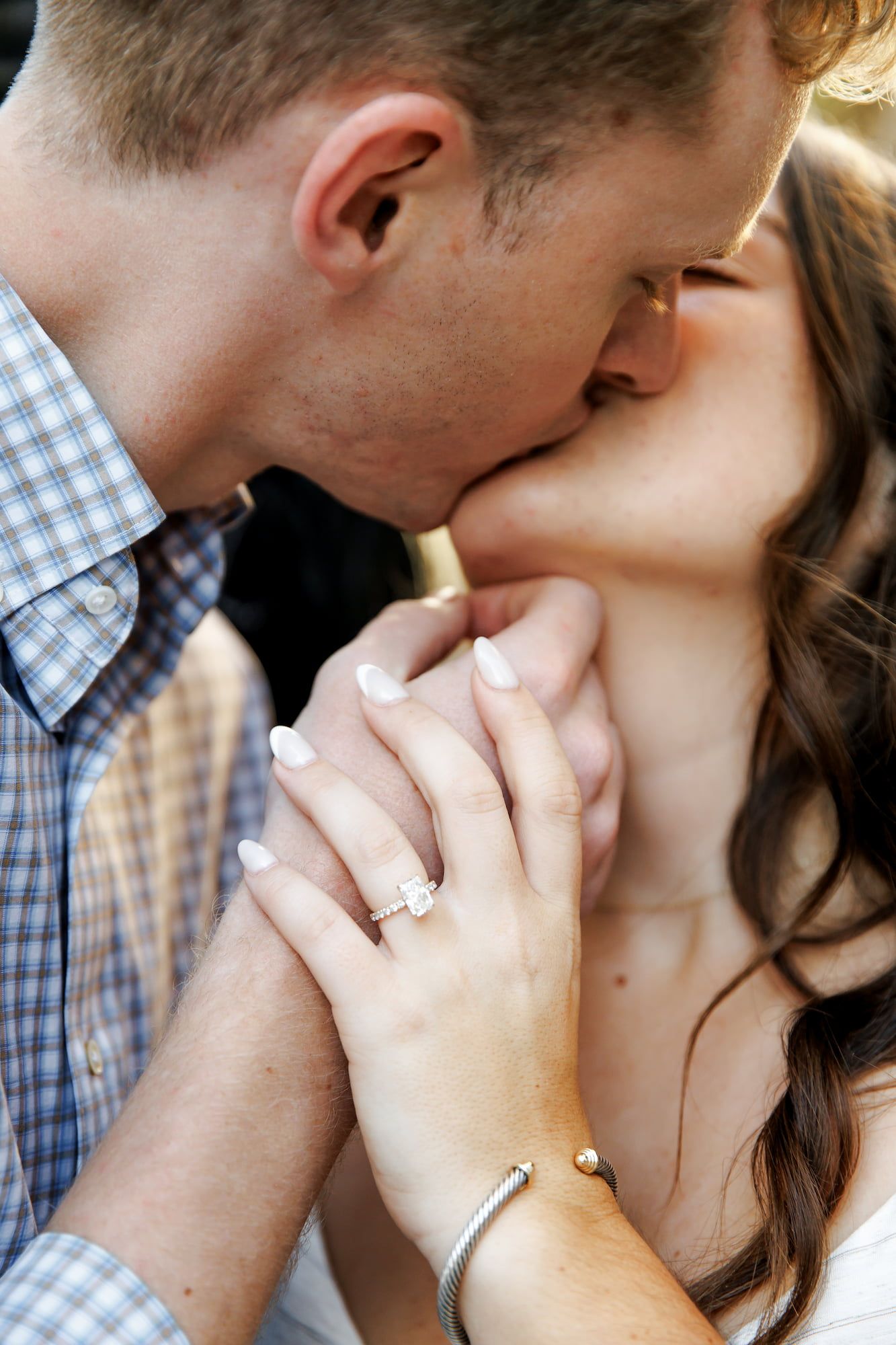 Couple sharing a romantic kiss while holding hands, showcasing a sparkling engagement ring and silver bracelet.