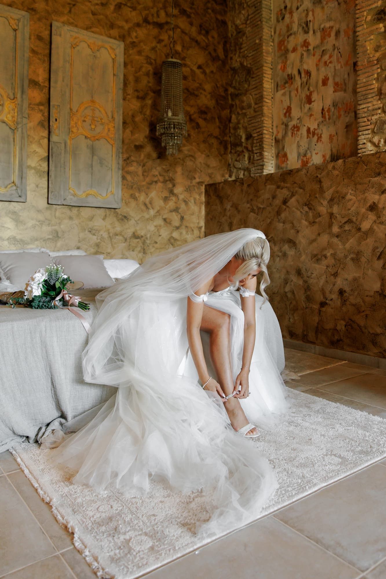 Bride in white gown and veil adjusting her heels while seated on a bed.