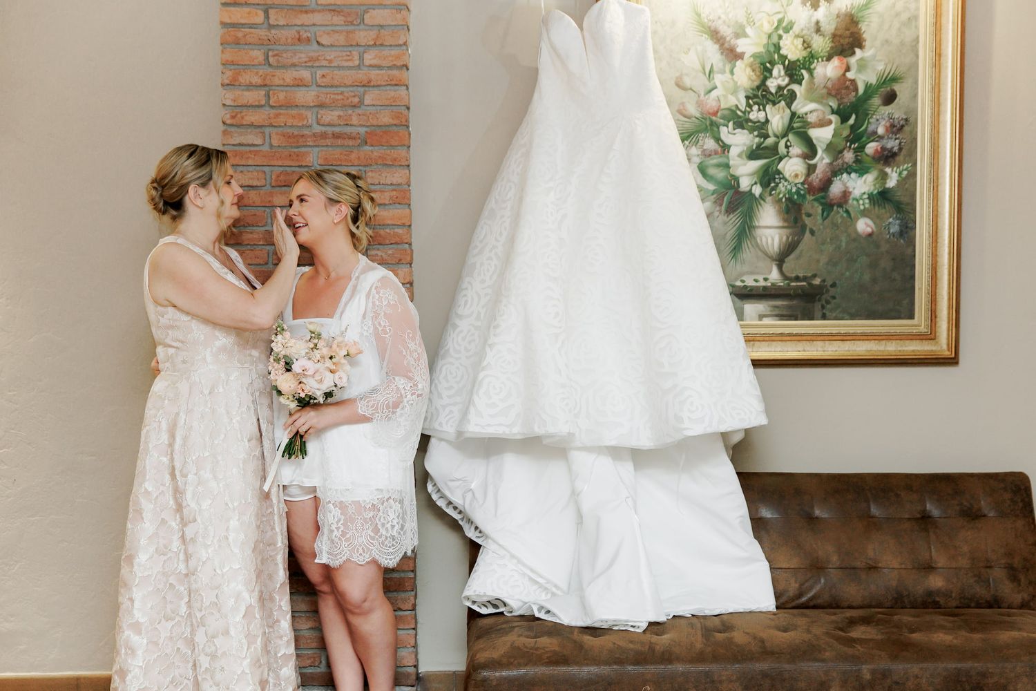 Bride and bridesmaid share a tender moment holding bouquet near hanging wedding gown and floral painting.