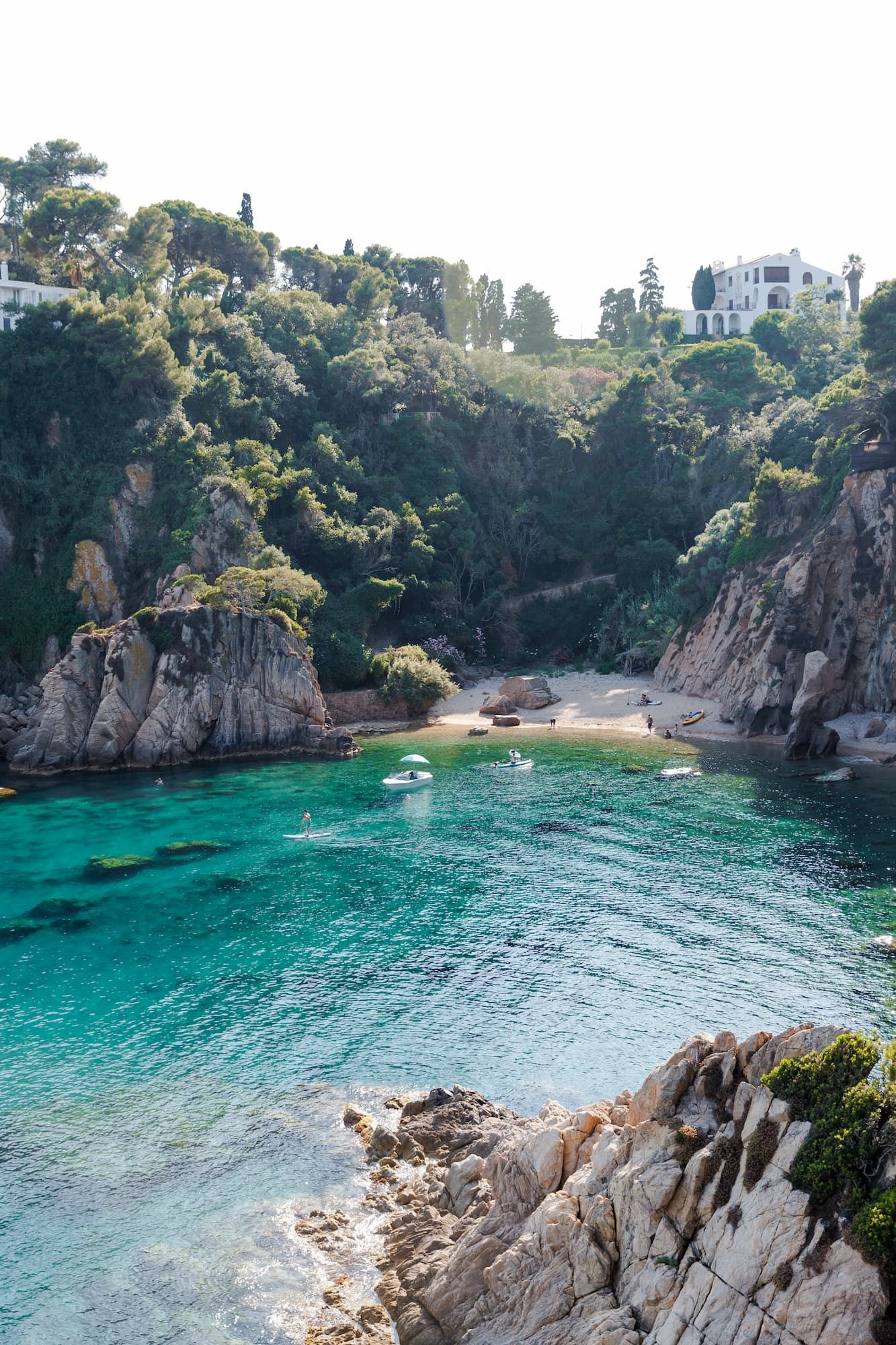 Turquoise Mediterranean cove with rocky cliffs, lush green trees, small sandy beach, and white villa overlooking crystal clear waters.
