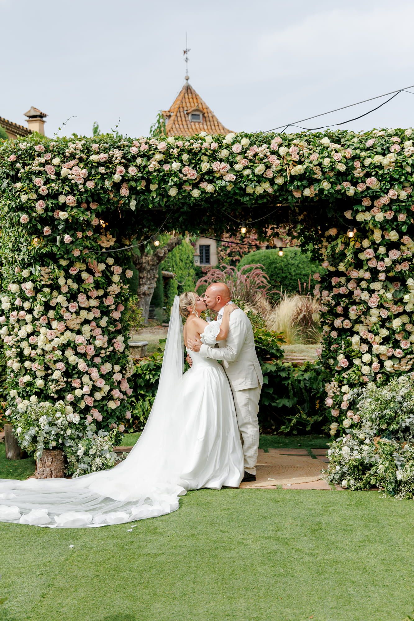 Bride and groom kissing under a floral arch covered in pink roses at an outdoor wedding ceremony.