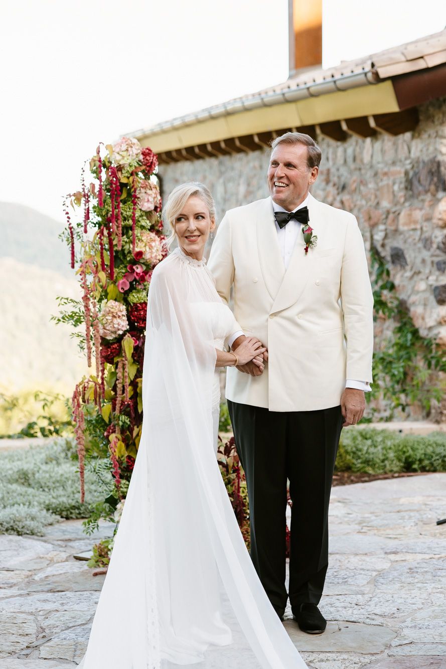Smiling bride in a flowing white gown and cape stands beside a groom in a white dinner jacket near a floral arch at a stone venue.
