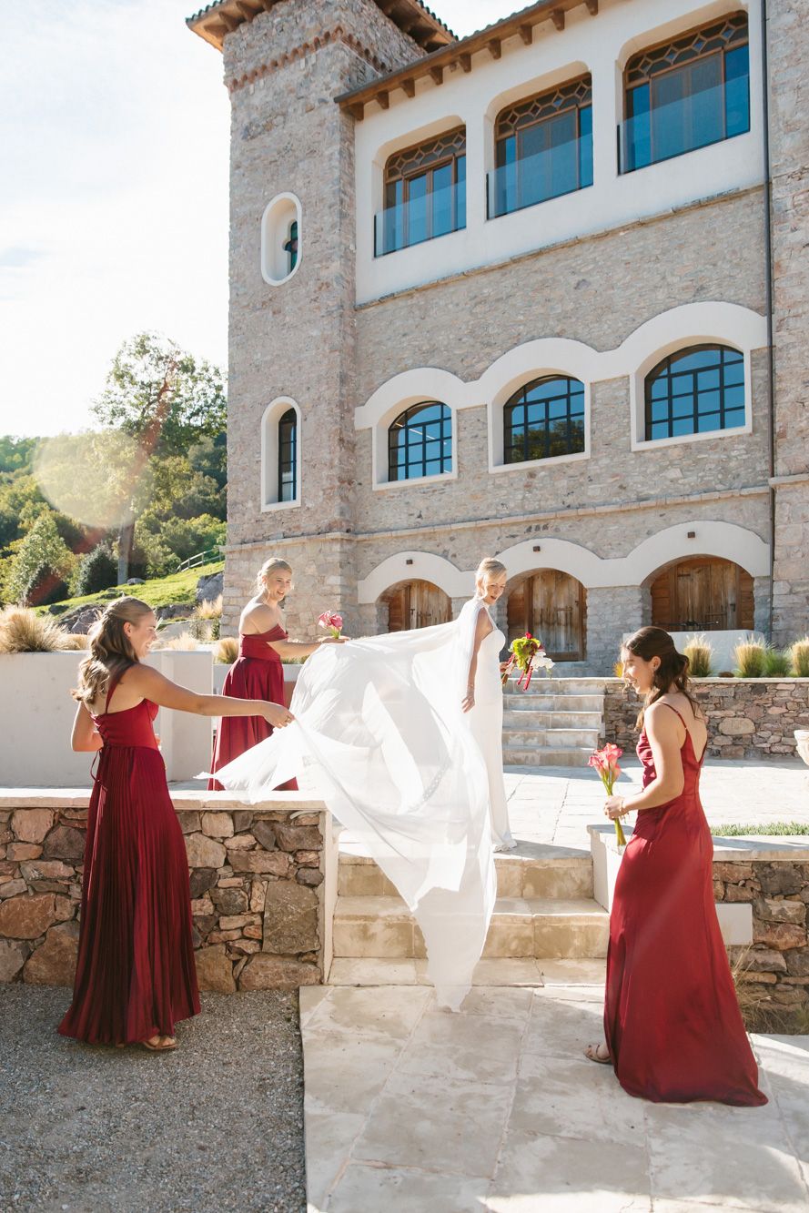 Bridesmaids in red dresses hold a flowing white wedding veil on stone steps outside a rustic stone venue with arched windows.