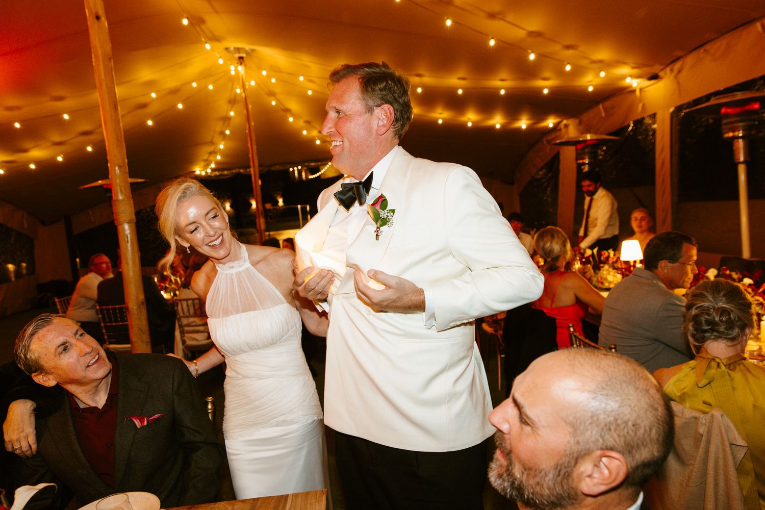 A groom in a white dinner jacket and bow tie laughs with a bride in a white halter gown at a lively tented wedding reception with string lig
