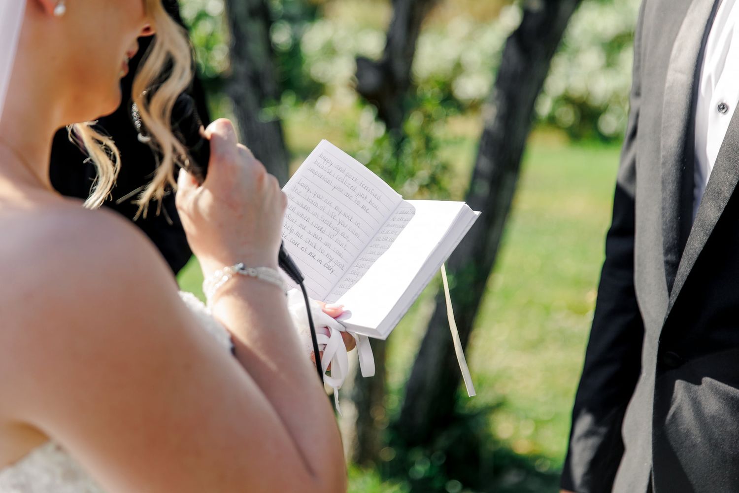 A bride reads her vow from a small book during an outdoor wedding ceremony in a garden setting.