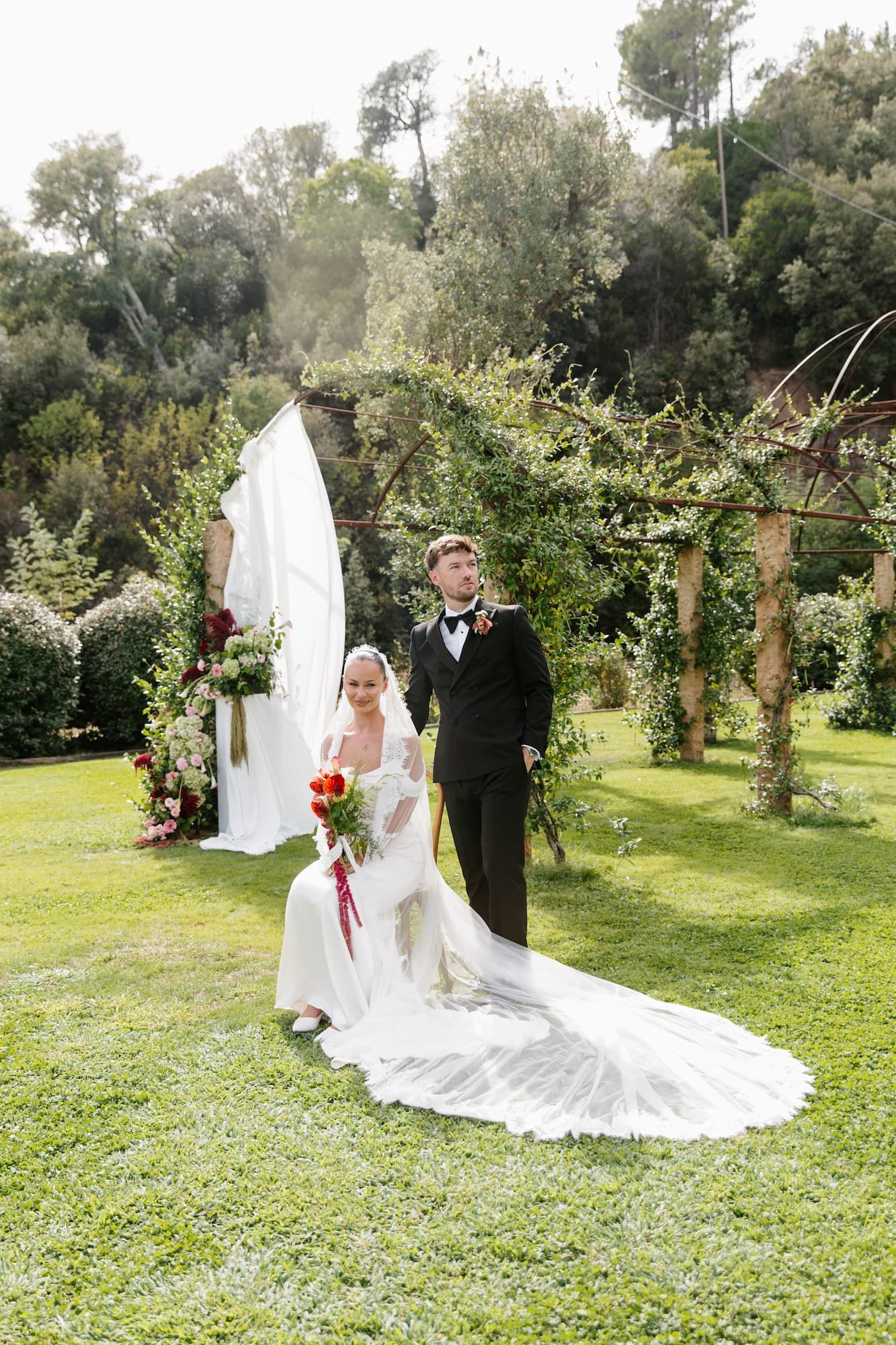 Bride in white gown with long train sits beside groom in black tuxedo at outdoor wedding ceremony with floral arch.