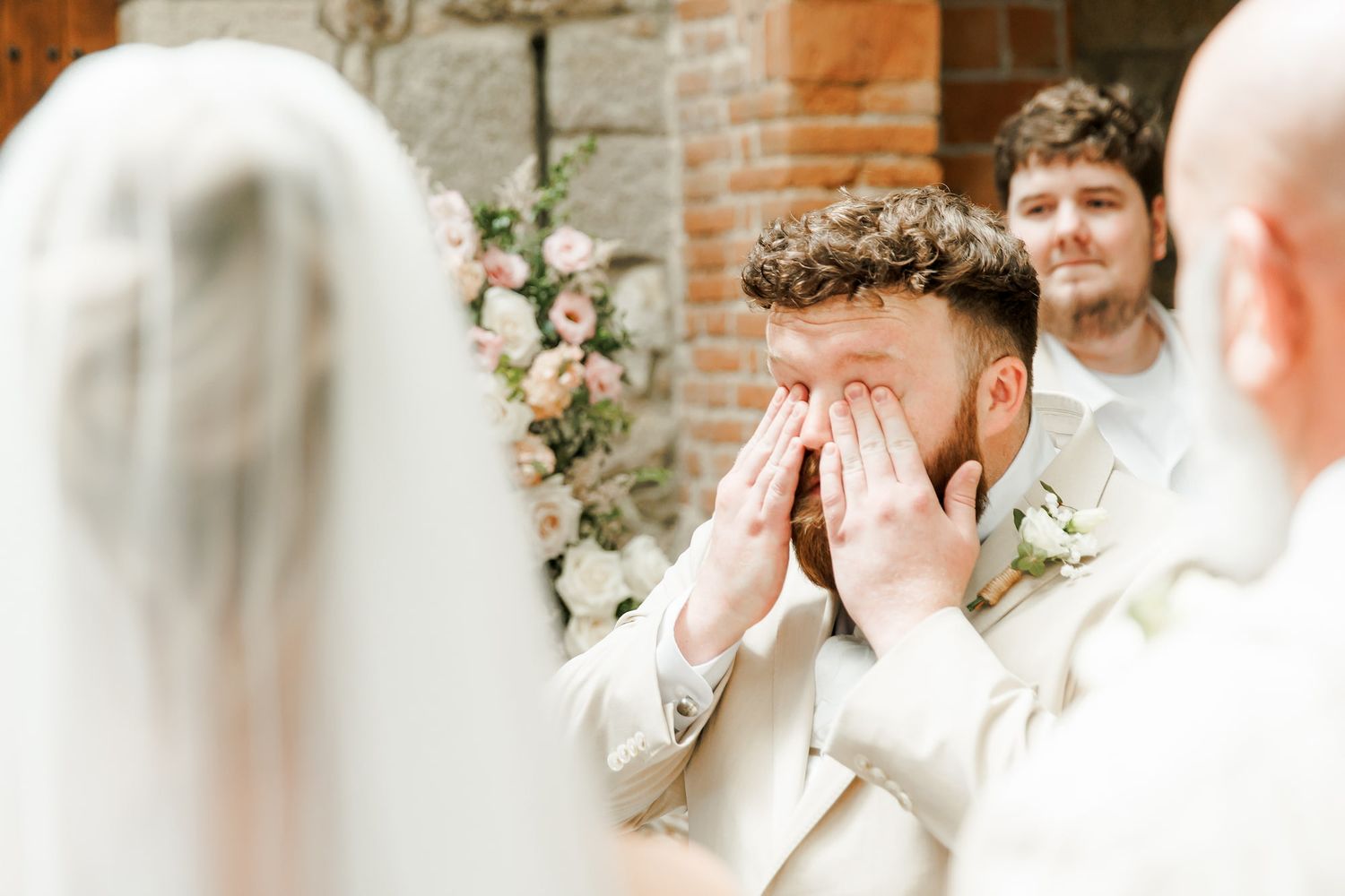 Emotional groom covers his face with hands as bride approaches at outdoor wedding ceremony with floral backdrop.
