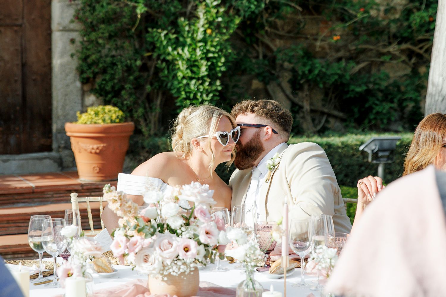 Bride and groom sharing a kiss at outdoor wedding reception table with floral centerpieces and wine glasses.