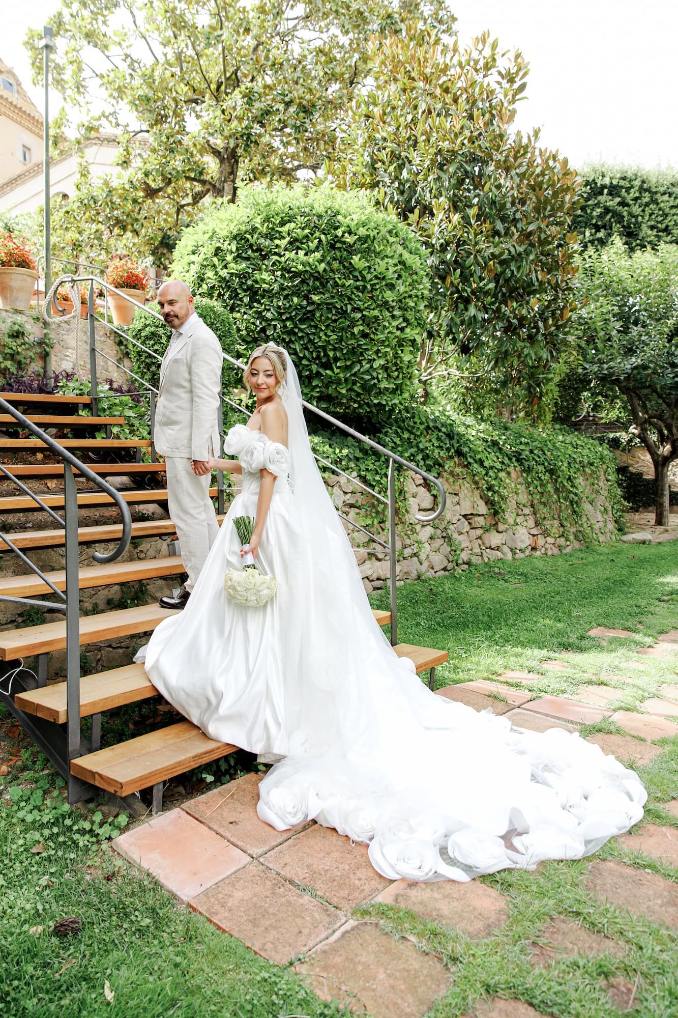 Bride in white gown with long train and groom in beige suit descend outdoor stone steps in lush garden setting.