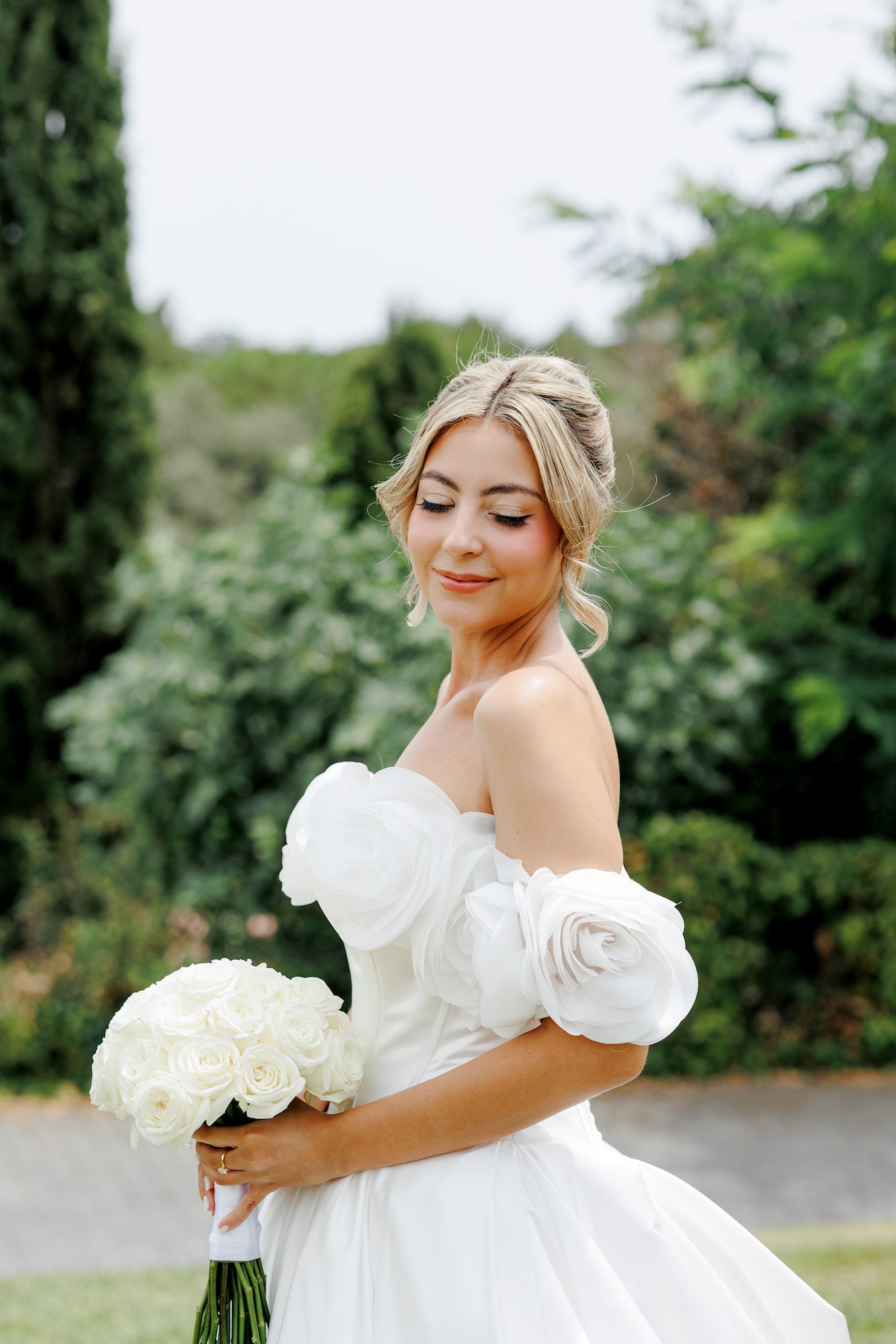 Bride in white off-shoulder gown with floral sleeves holds white rose bouquet outdoors among lush green trees.