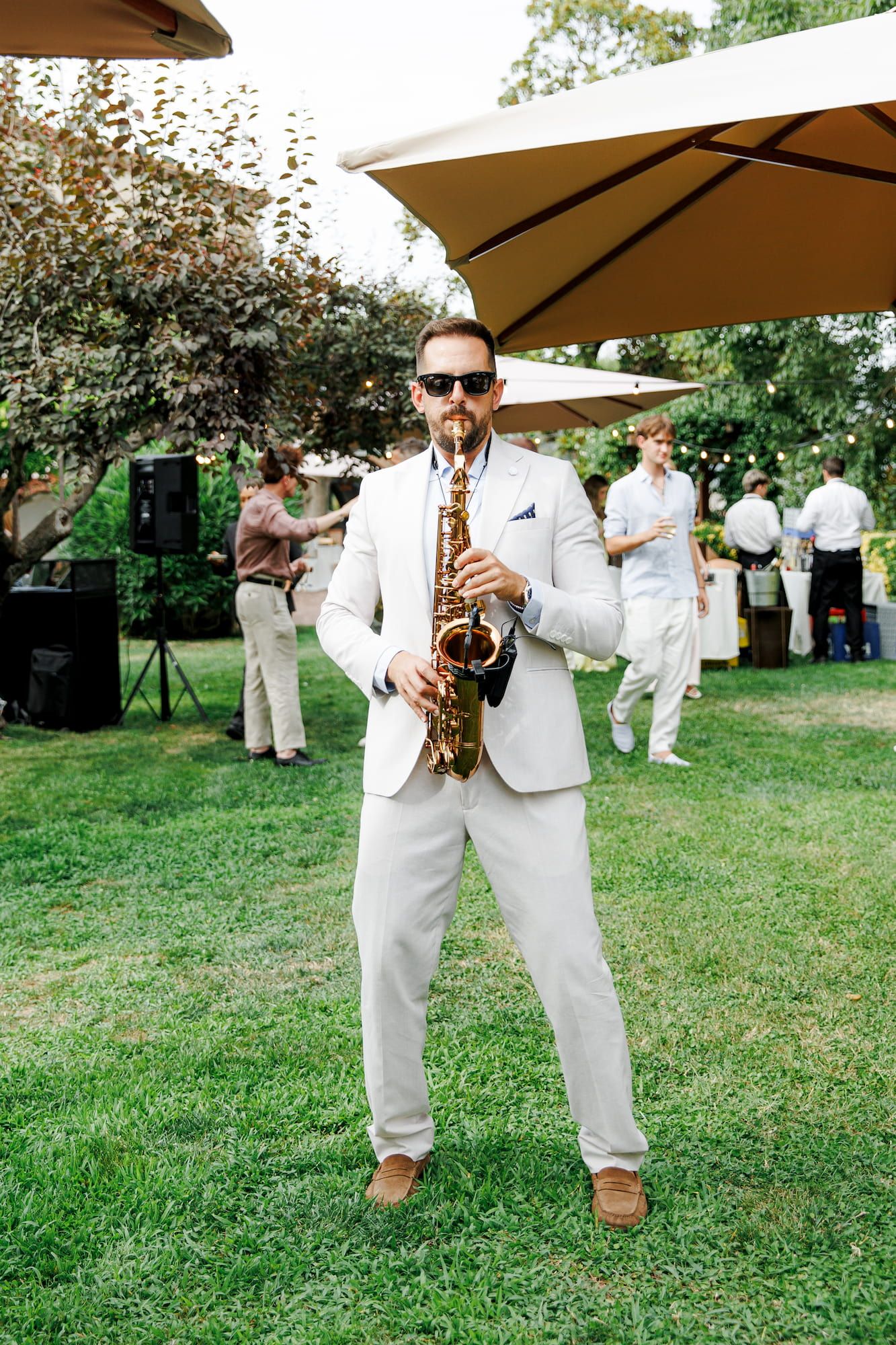 Musician in a white suit playing saxophone at an outdoor wedding reception on a sunny day.
