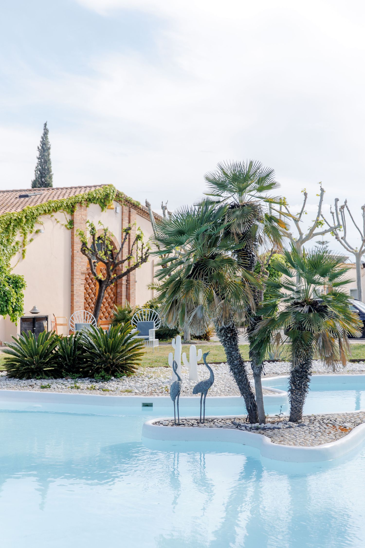 Elegant pool with palm trees, crane sculptures, and a stone villa surrounded by lush greenery under a cloudy sky.