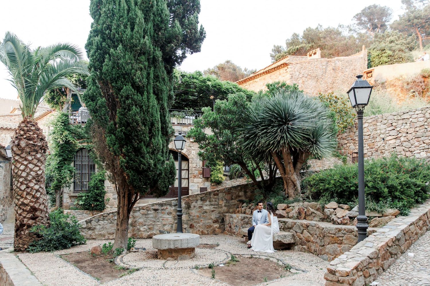 Stone pathway winding through a Mediterranean garden with palm trees and rustic walls in a tranquil setting.