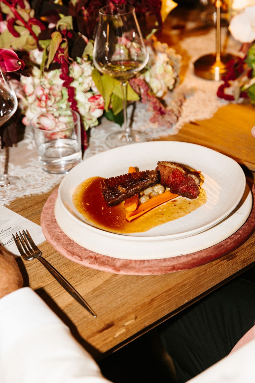 Gourmet plated entrée with sliced seared meat, glazed carrots and golden sauce on a white plate, set on an elegant floral table.