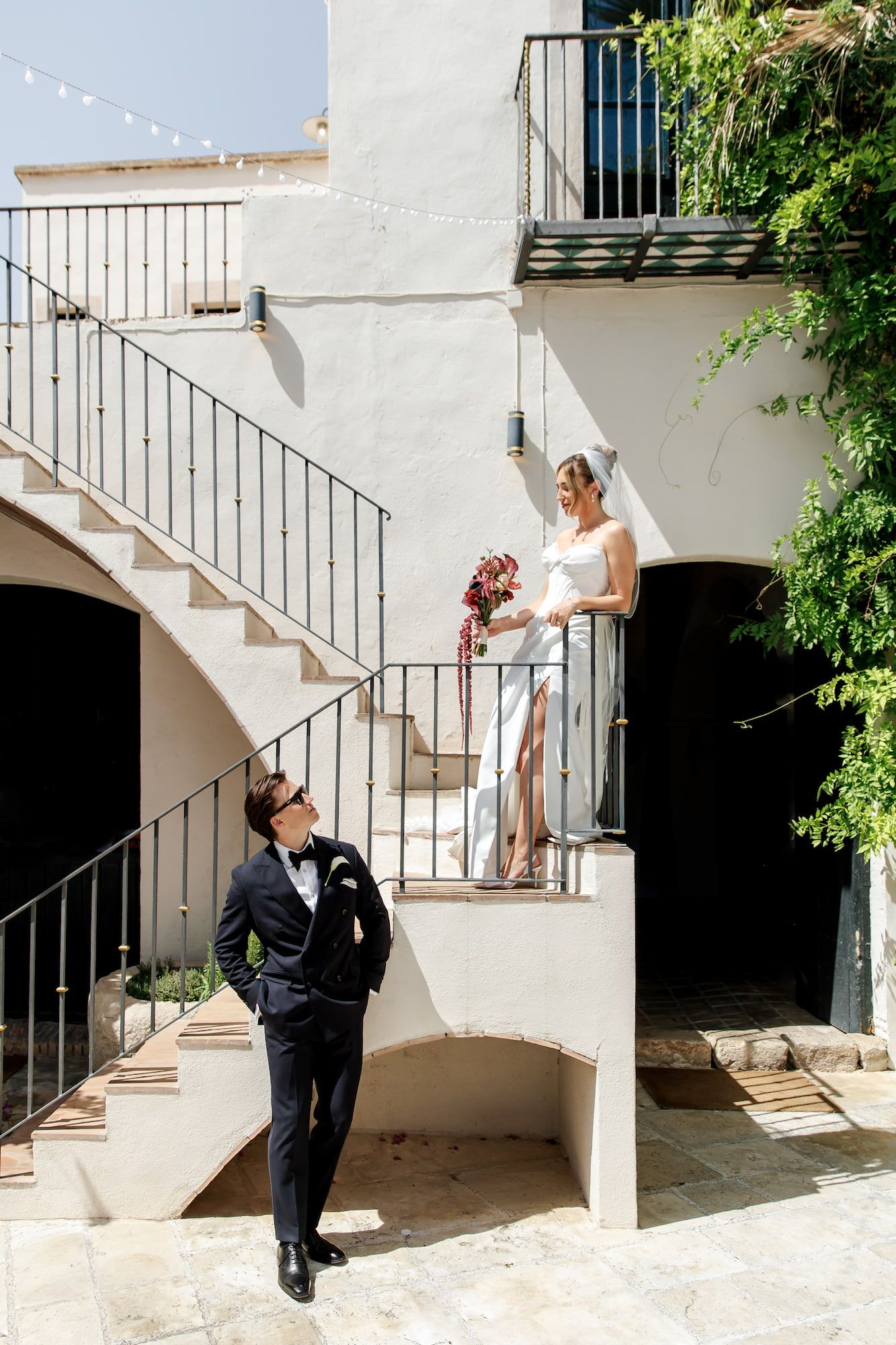 Bride in a white gown stands on the staircase landing as the groom in a navy tuxedo looks up at her at an outdoor wedding venue in Barcelona
