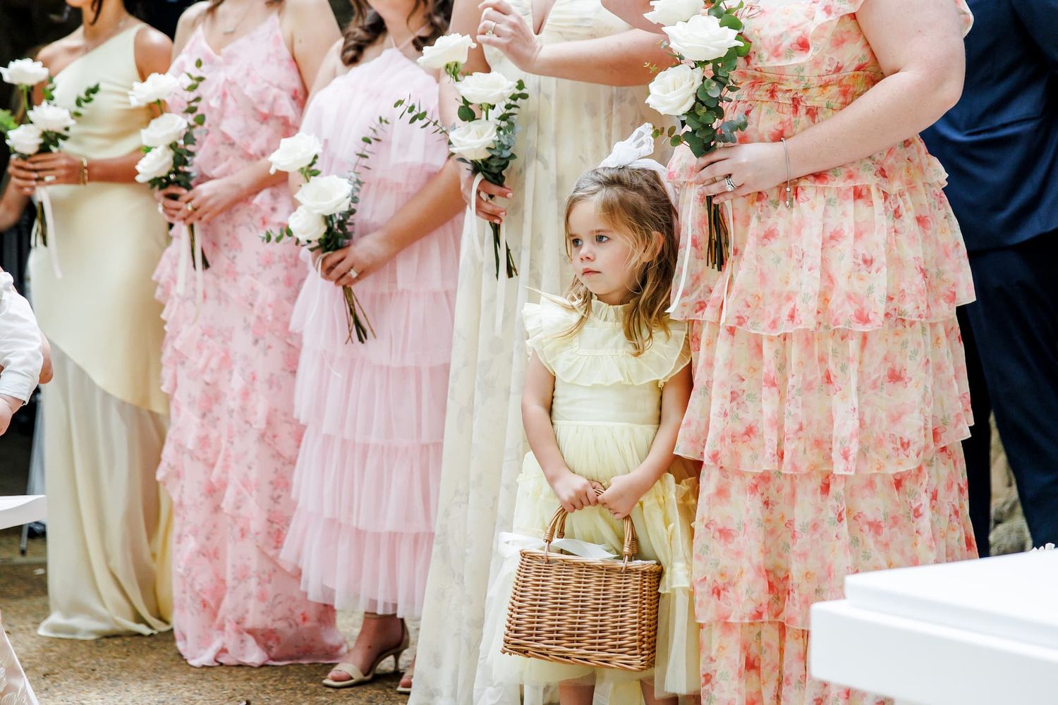 A young flower girl in yellow dress holds wicker basket amid bridesmaids in pink floral gowns at outdoor wedding.