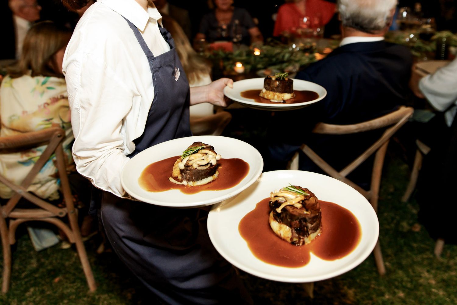 Waiter serving elegant plated dishes with rich brown sauce at a formal outdoor dining event.