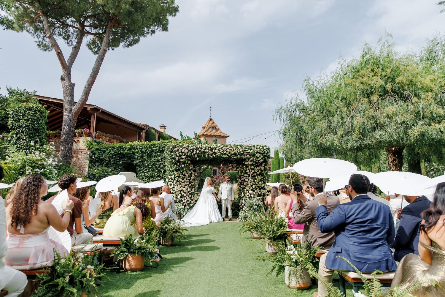 Outdoor wedding ceremony on green lawn with floral arch, guests seated with white umbrellas, surrounded by lush greenery at weddind venue.