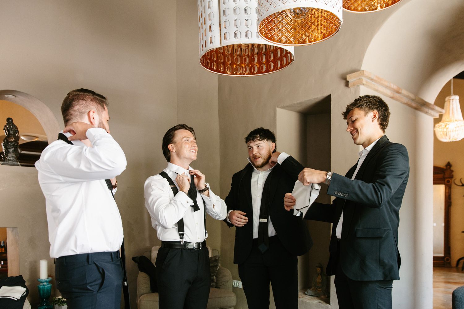 Four groomsmen in formal attire adjusting ties and cufflinks while getting ready indoors under warm pendant lights.