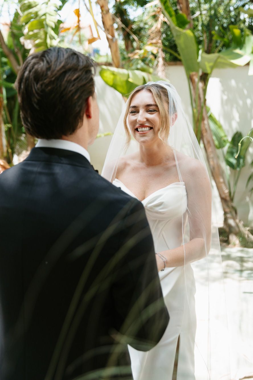 Smiling bride in white strapless gown and veil exchanges vows with groom in black suit outdoors.