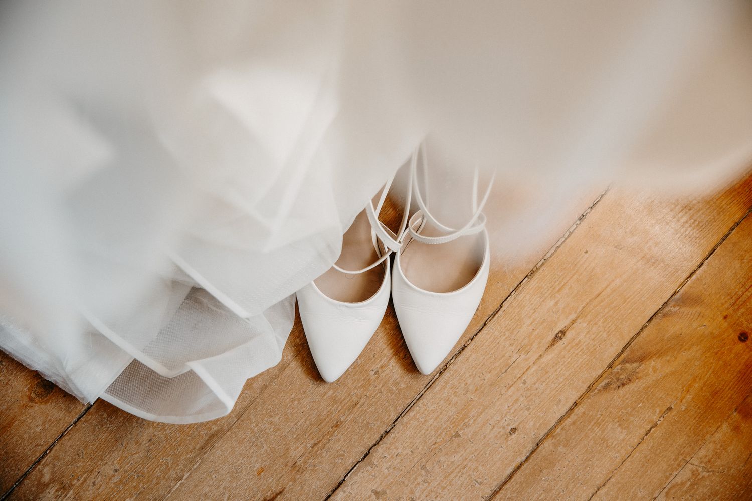 White pointed-toe bridal heels with ankle straps resting on wooden floor beside flowing white wedding dress.