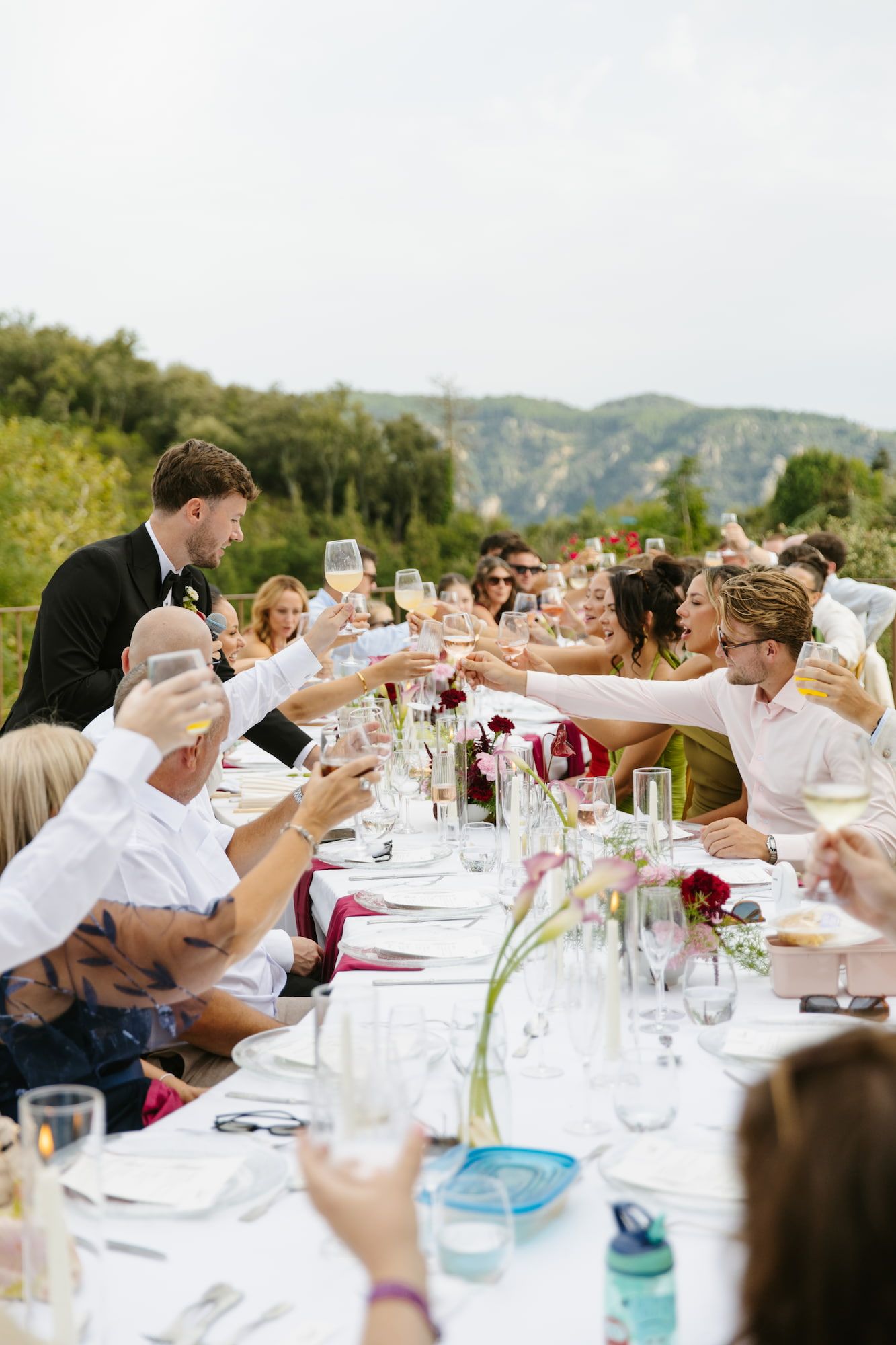 Outdoor wedding reception toast with guests raising wine glasses along a long decorated table with mountain views.
