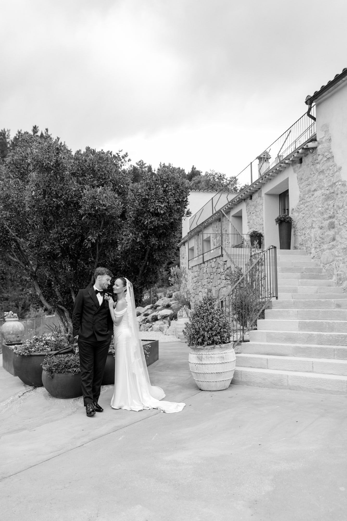 Black and white photo of bride and groom sharing a romantic moment outside a stone villa with lush gardens.