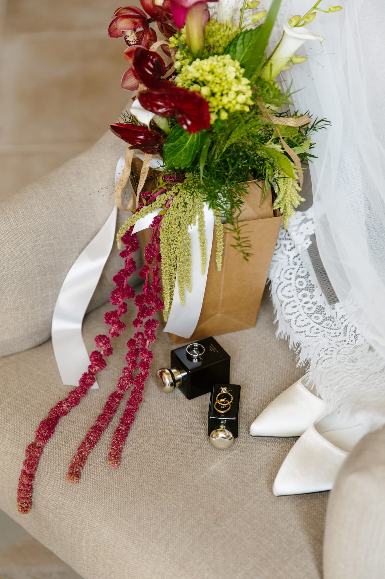 Bridal flat lay with bouquet, white heels, perfume bottles, and rings on a beige chair.