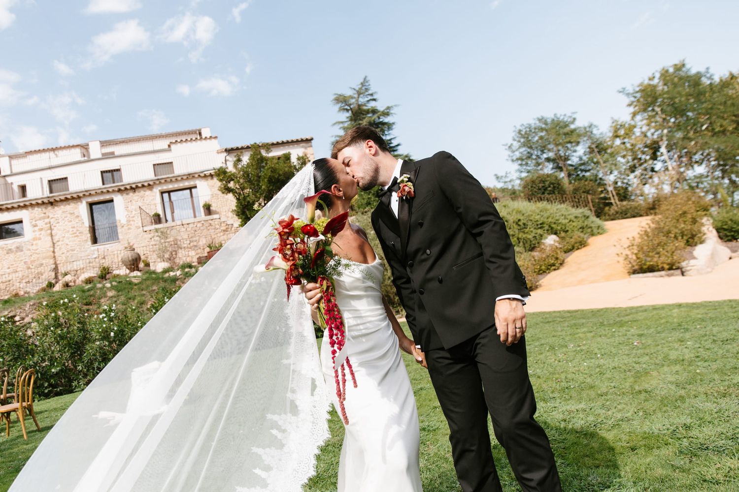 Bride and groom share a romantic kiss outdoors at a vineyard wedding venue, with a flowing veil and red bouquet.