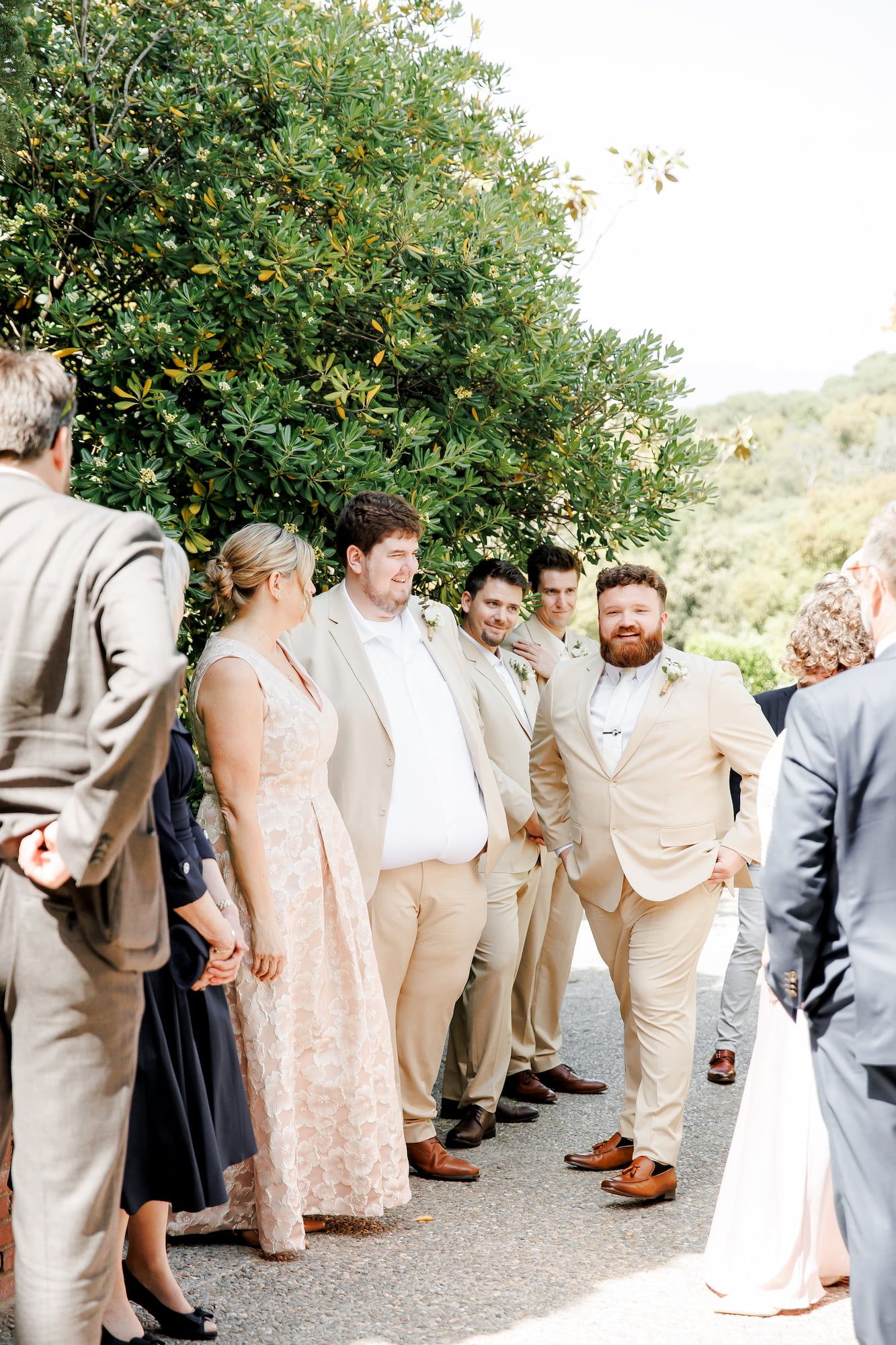 Groomsmen in tan suits gather outdoors with wedding guests near lush green trees before the ceremony.