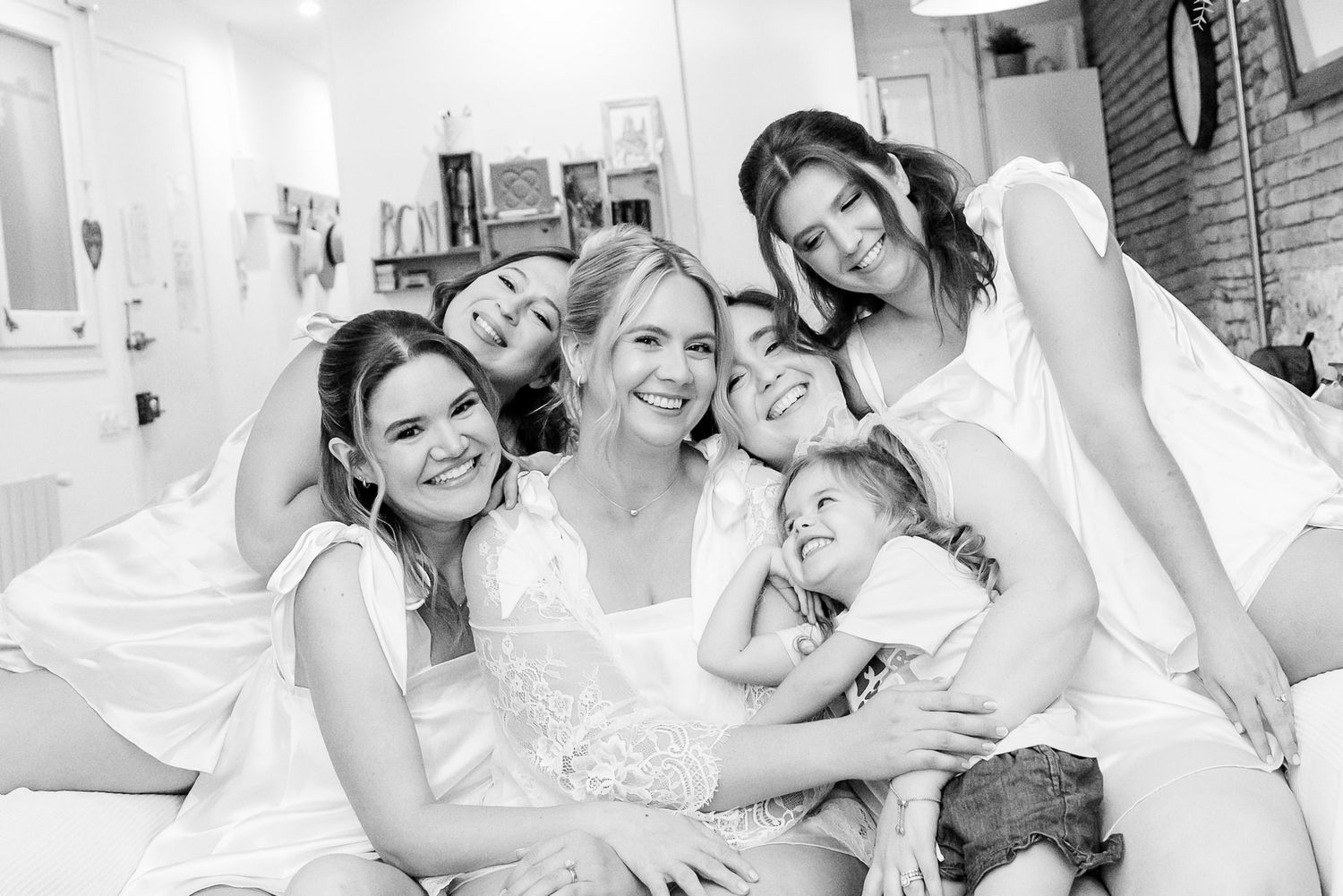 Black and white photo of a bride surrounded by smiling bridesmaids and a child, posing together on a couch.