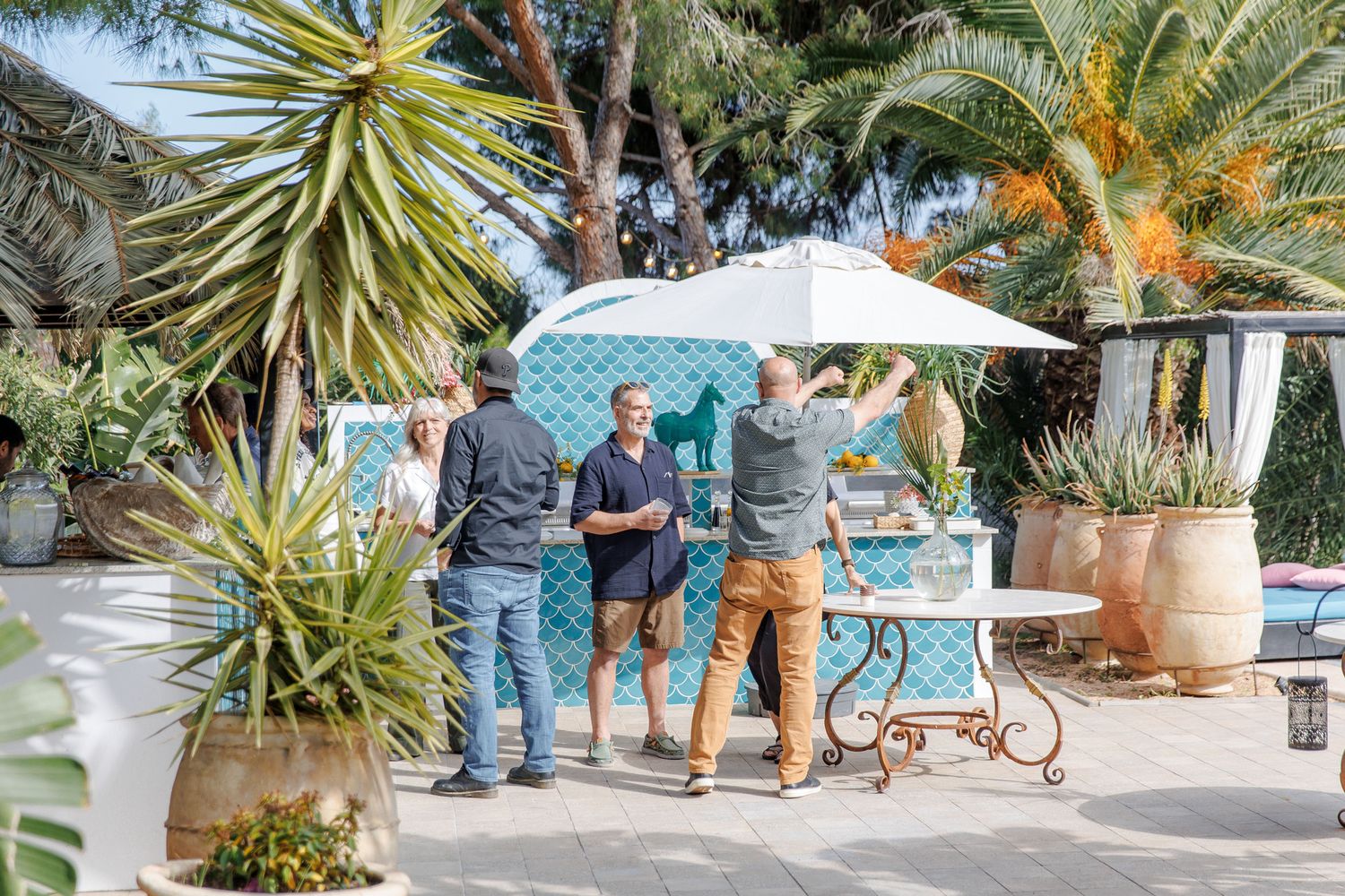 Group of people socializing outdoors in a lush garden patio with tropical plants and a white umbrella.