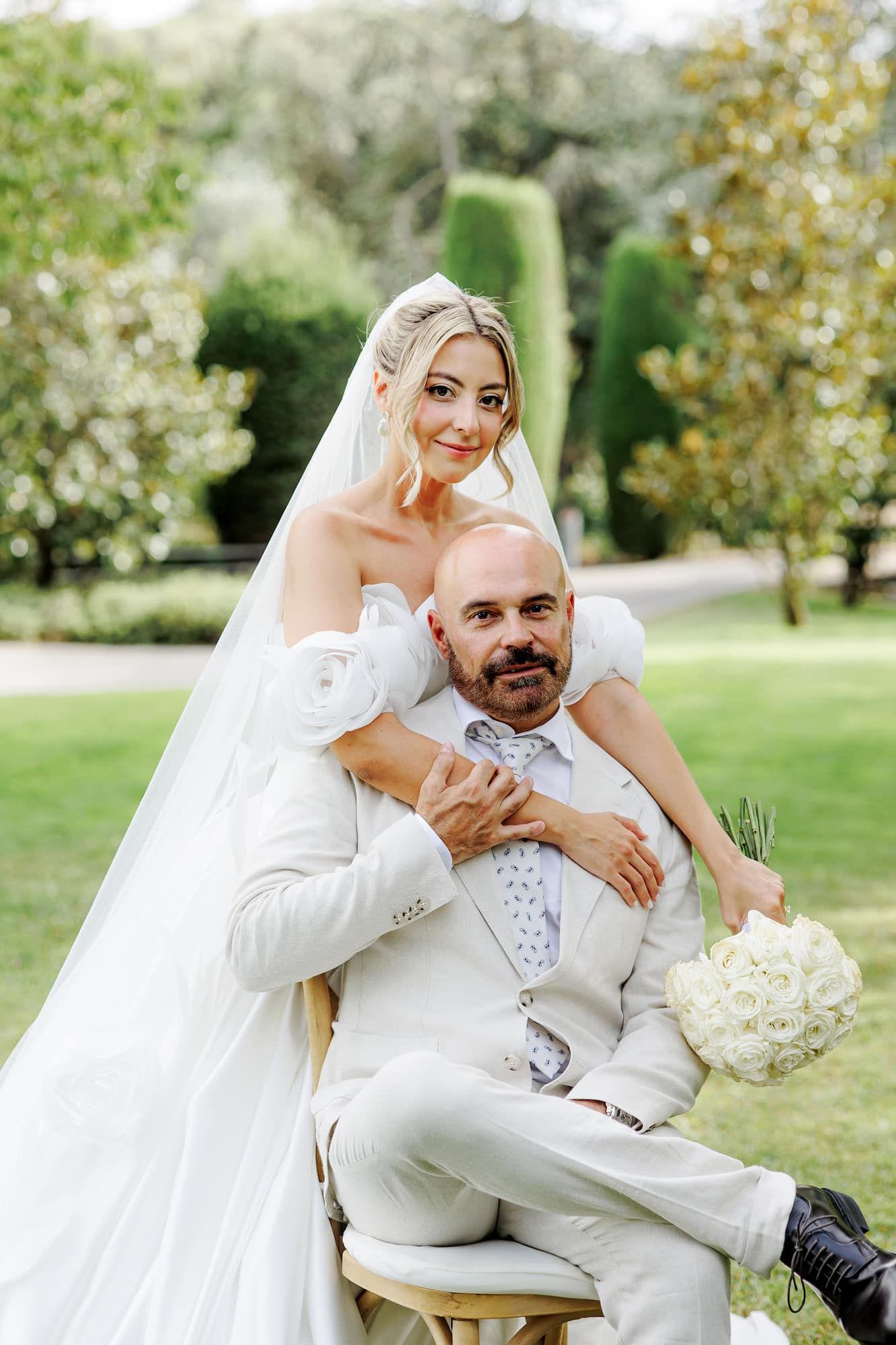 Bride in white gown embraces groom in light suit seated outdoors, holding white bouquet in lush garden.