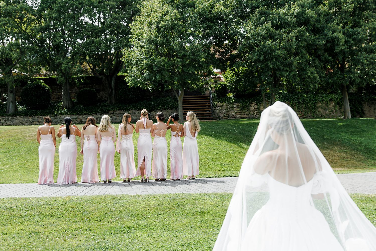 Bride in white gown and veil approaches bridesmaids in pink dresses standing on green lawn at outdoor wedding.