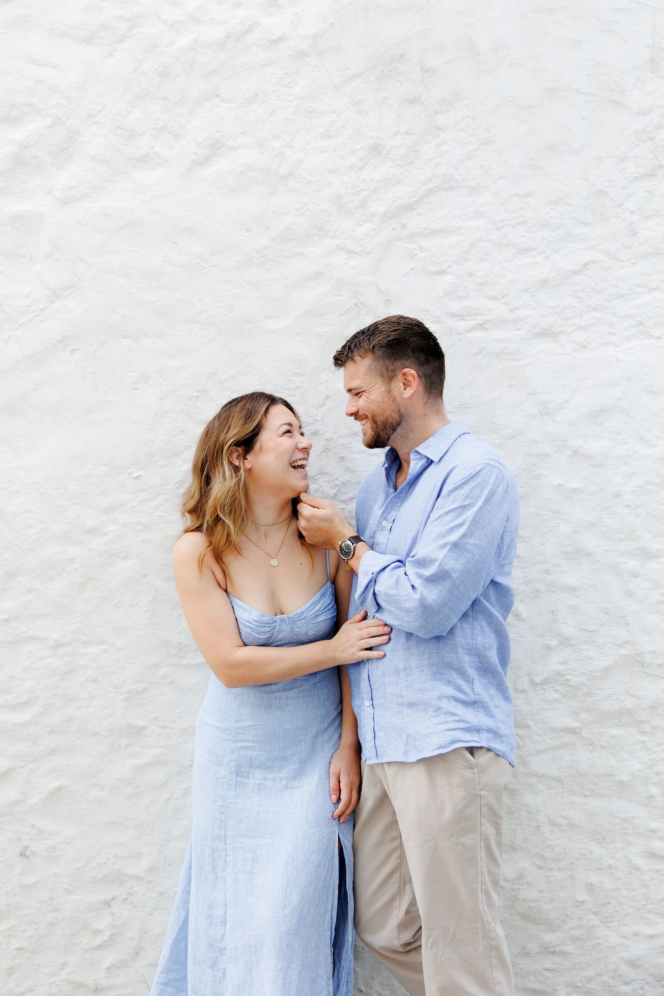 A couple in coordinating light blue outfits share intimate laughs against a white wall in a romantic photo series.