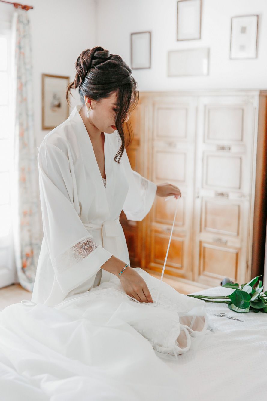 Bride in white robe sitting on bed holding wedding dress ribbon in bright bedroom.