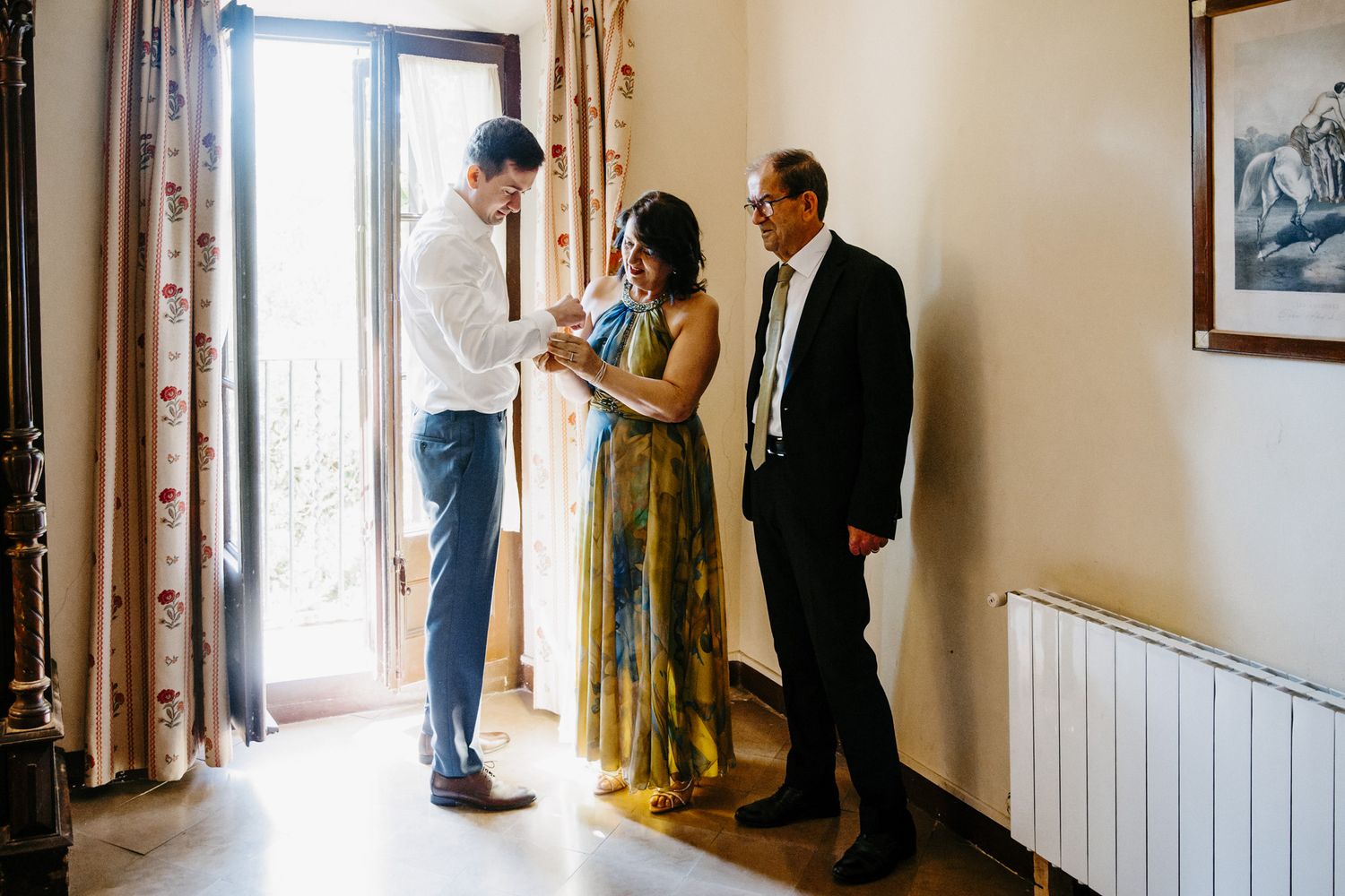 Three elegantly dressed people share a warm moment indoors near a sunlit window during a wedding celebration.